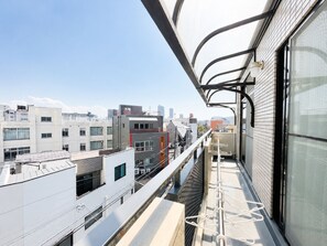 A balcony with a view of city buildings, a metal railing, and a window with a white curtain. - SERENE | Nishikujo, 5th Floor | 100 ㎡, Sleeps 10 | Direct Access to USJ and Osaka Station (Konohana Ward)