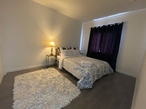 A bedroom with a bed, a nightstand, a lamp, a large fluffy rug, and a window with curtains.