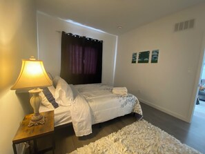 Um quarto com uma cama, uma mesa de cabeceira com abajur, uma janela com cortinas e um tapete pequeno.