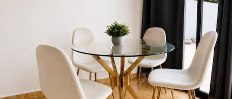 Un comedor con una mesa de vidrio y cuatro sillas blancas, una planta pequeña en maceta y vistas al exterior a través de una ventana con cortinas negras.