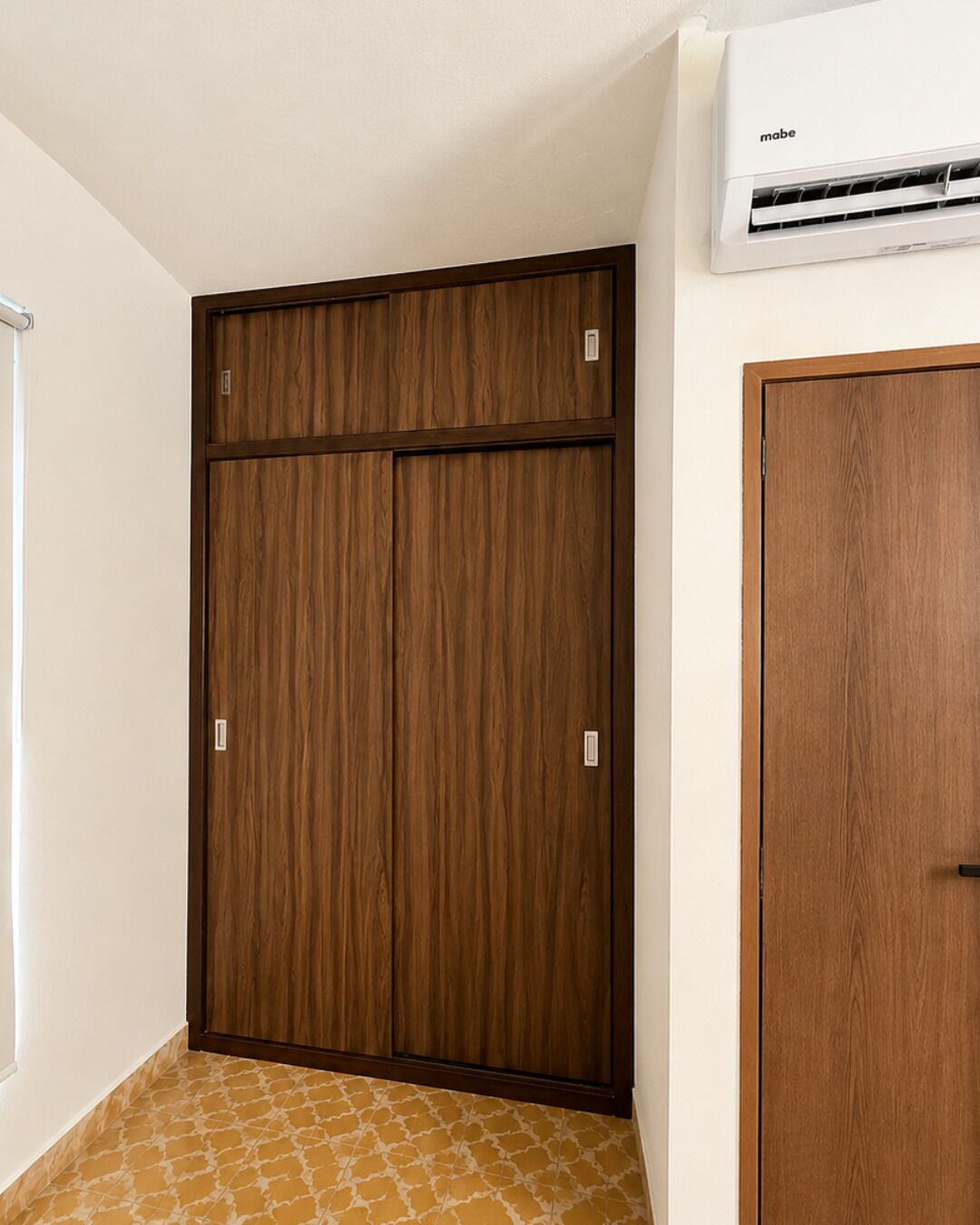 A room with a wooden wardrobe, a smaller wooden cabinet, and a wall-mounted air conditioning unit.
