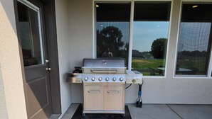 En gasolgrill med fyra brännare och ett sidoskåp, placerad i ett takförsett utomhusområde med utsikt över en gräsyta genom stora fönster.