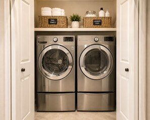 A laundry room with two stainless steel front-loading washers and dryers, a shelf above with baskets labeled 'POOL TOWELS' and 'LAUNDRY PODS,' and a small potted plant.