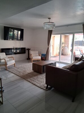 A modern living room with a flat-screen TV, a chandelier, a sofa, a coffee table, and a large sliding glass door leading to an outdoor area.