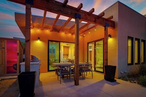 A covered outdoor patio with a dining table and chairs, a grill, and a sliding glass door leading to an interior kitchen. - Sagebrush Hideaway Pool, Sauna, Cold Plunge, Views (Pioneertown)