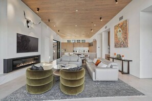 A modern living room with a fireplace, a large painting, and a kitchen in the background.