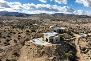 A modern two-story house with a pool, surrounded by a desert landscape and mountains.
