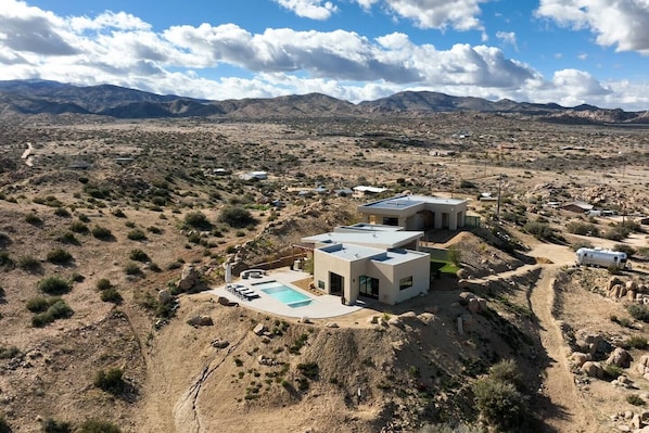 A modern two-story house with a pool, surrounded by a desert landscape and mountains.