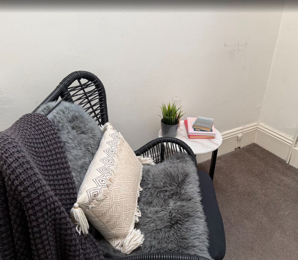A corner of a room with a black chair, a grey furry cushion, a patterned pillow, a small potted plant, and a stack of books on a white table.