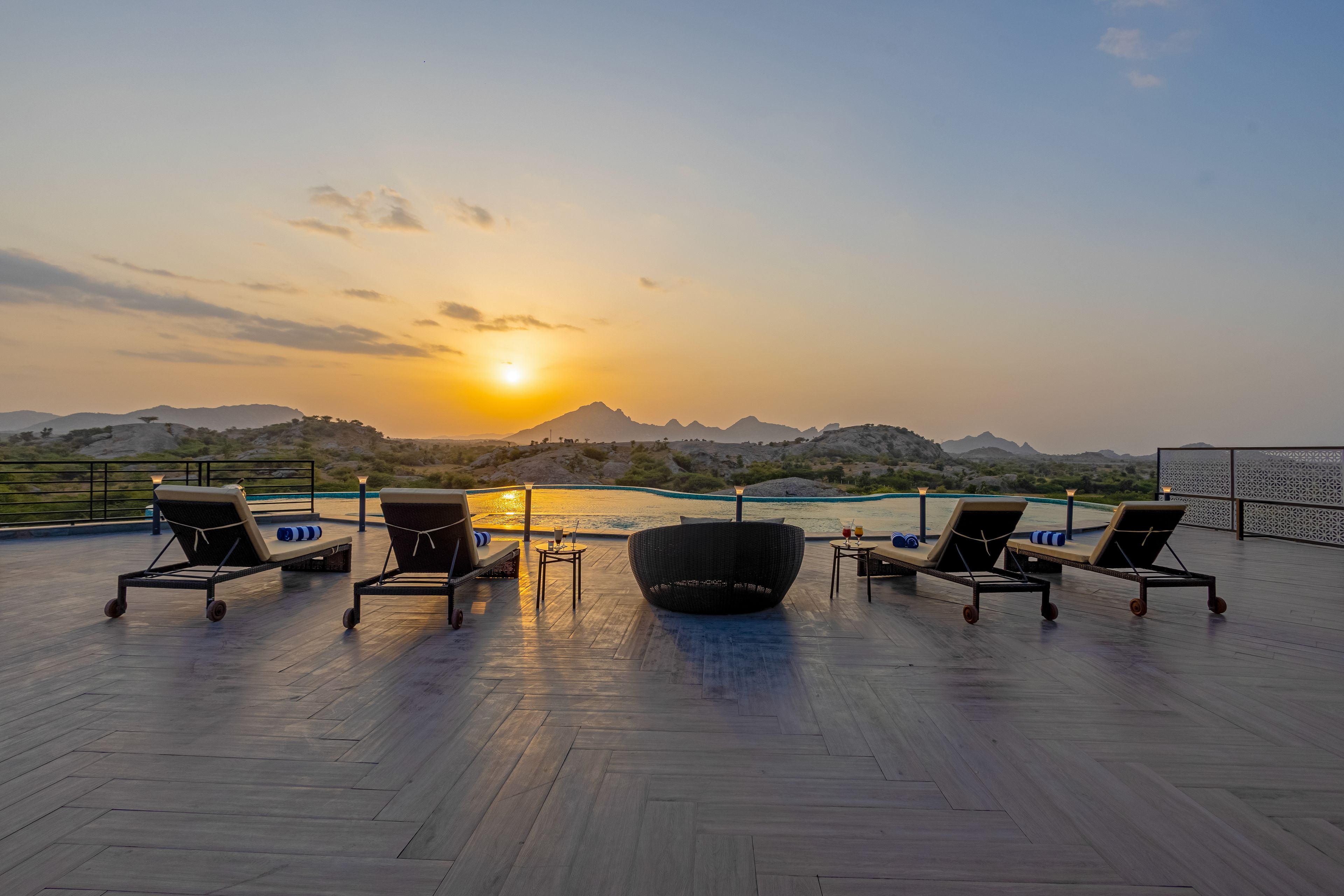 A rooftop terrace with lounge chairs, a table, and a view of the sunset over a body of water and mountains.