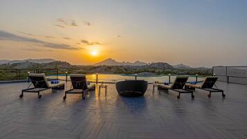 A rooftop terrace with lounge chairs, a table, and a view of the sunset over a body of water and mountains.