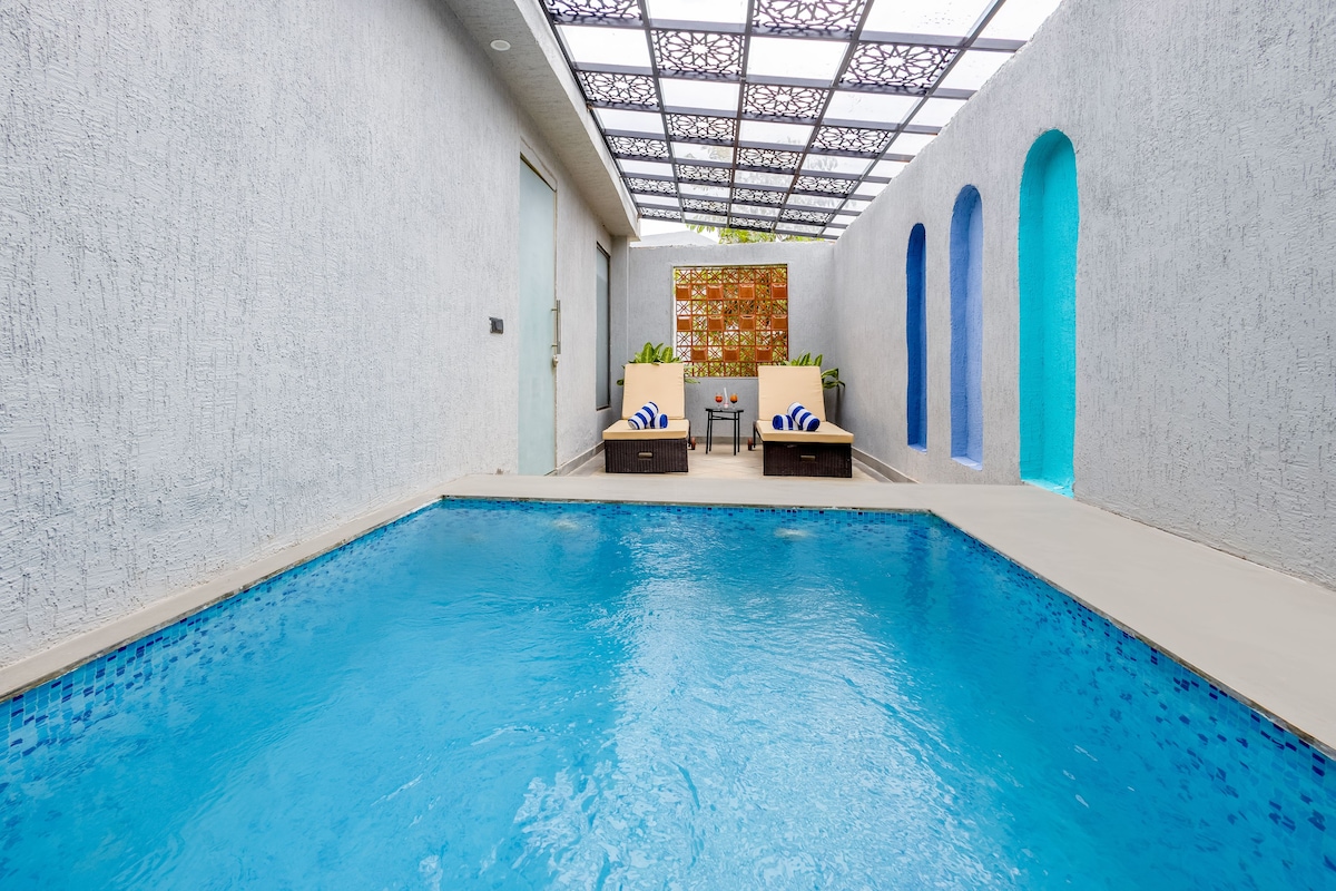 An indoor swimming pool with a clear view of the pool area, including the pool's edge, a seating area with chairs, and a decorative wall.