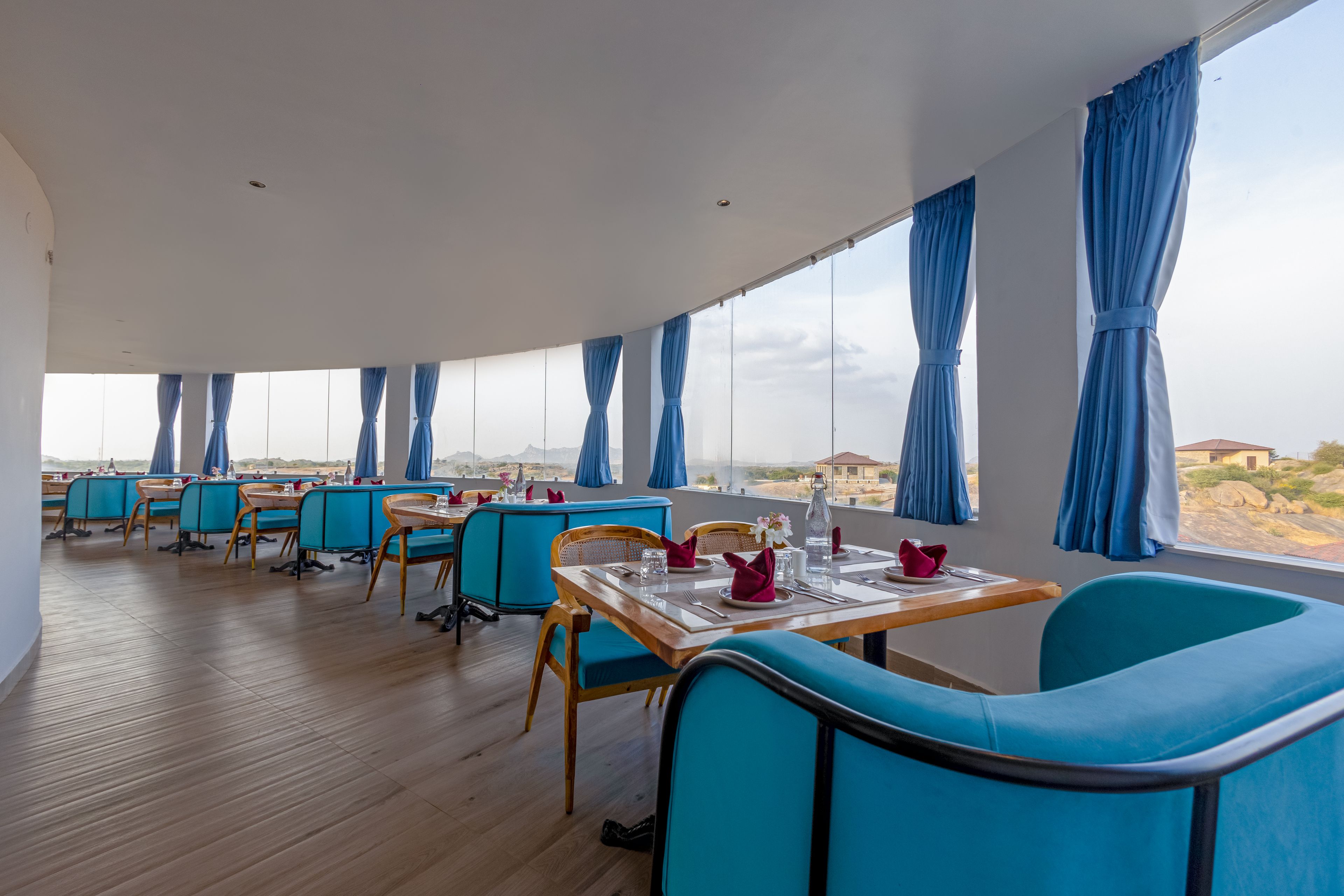 A dining area with tables and chairs, large windows with curtains, and a view of the landscape.