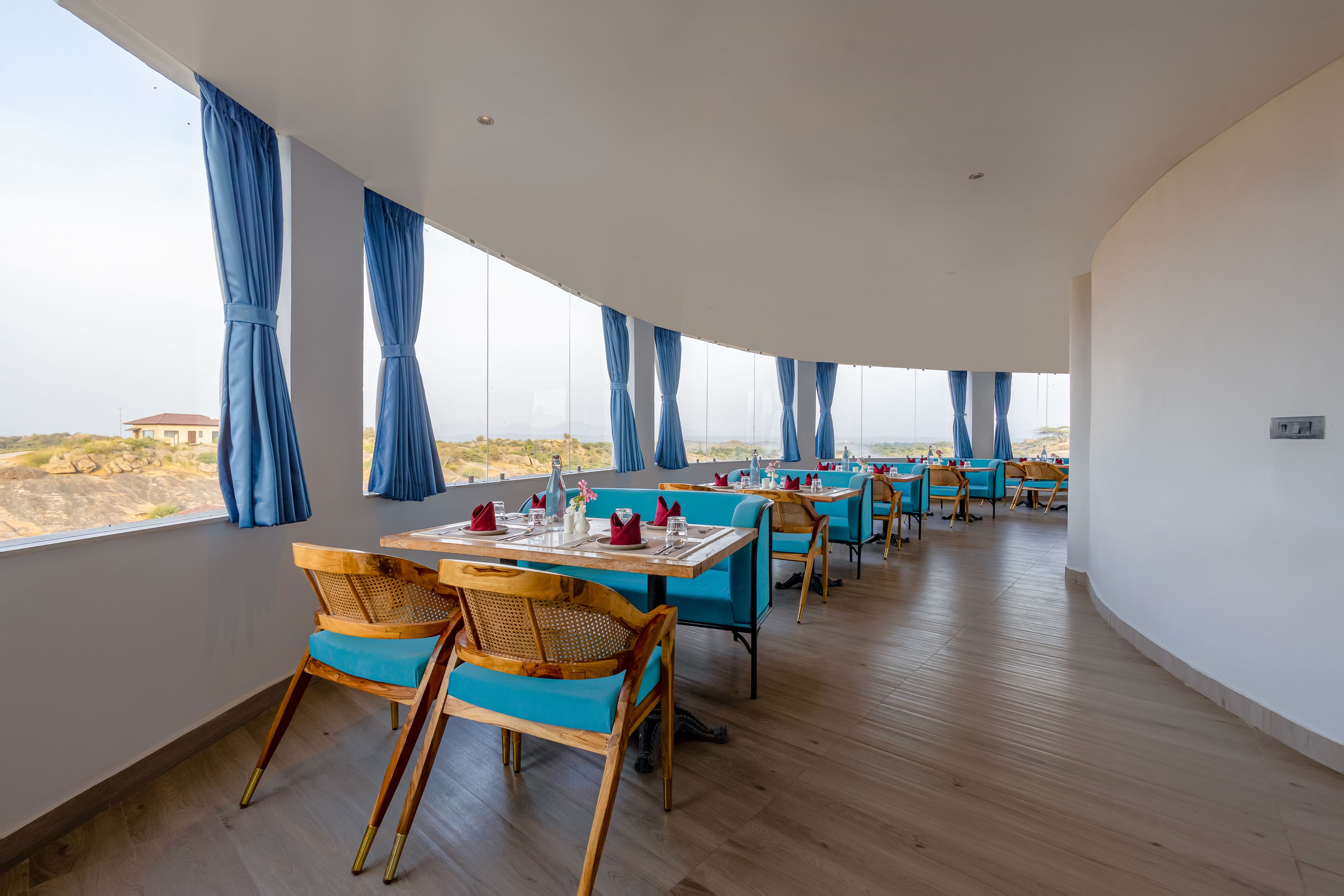 A restaurant with wooden chairs and tables, large windows with blue curtains, and a view of the landscape.