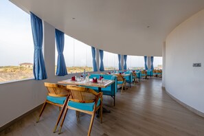 A restaurant with wooden chairs and tables, large windows with blue curtains, and a view of the landscape. - Sterling Leopard Haven Jawai (Bali)