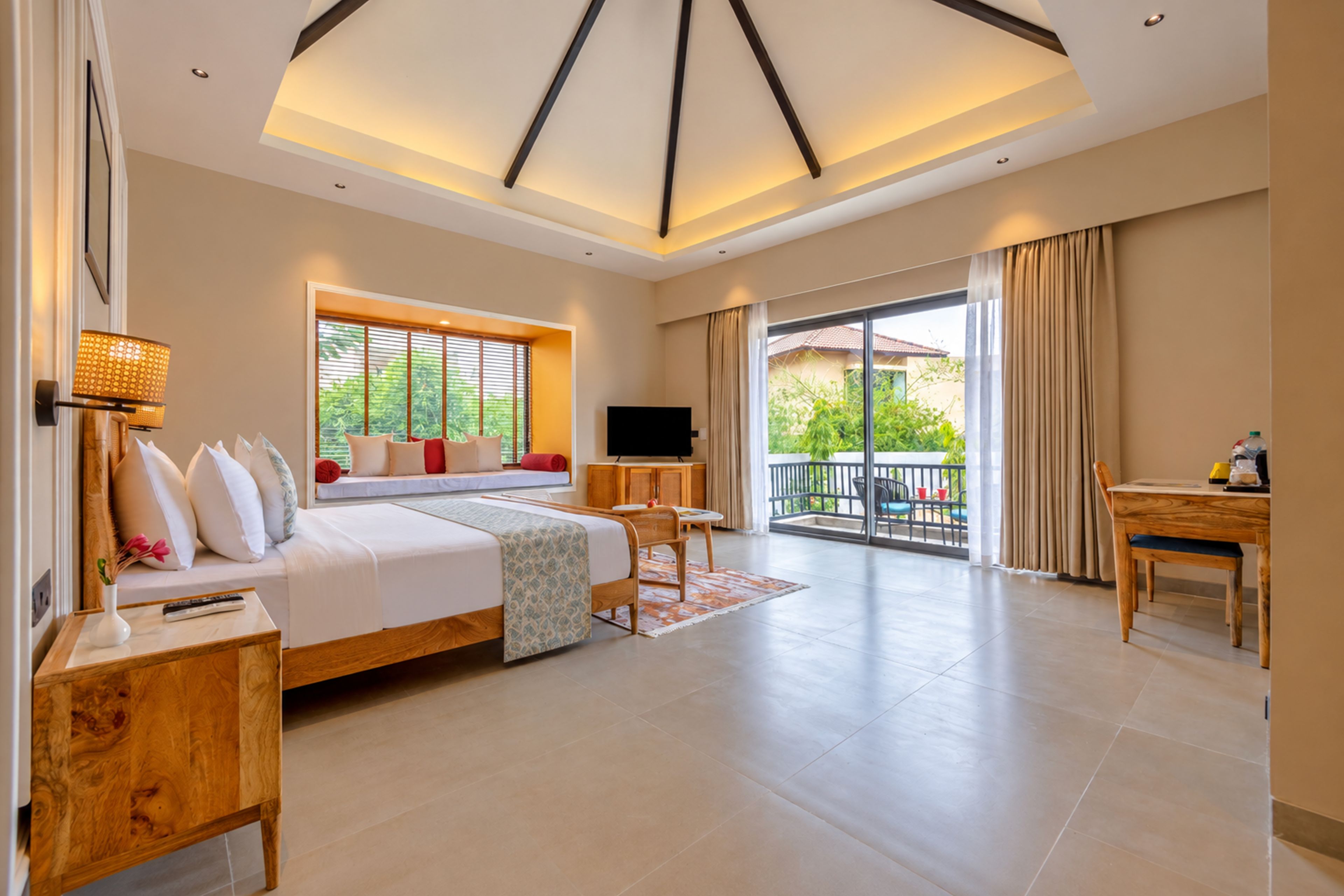 A hotel room with a bed, a TV, a desk, and a balcony with a view of greenery.