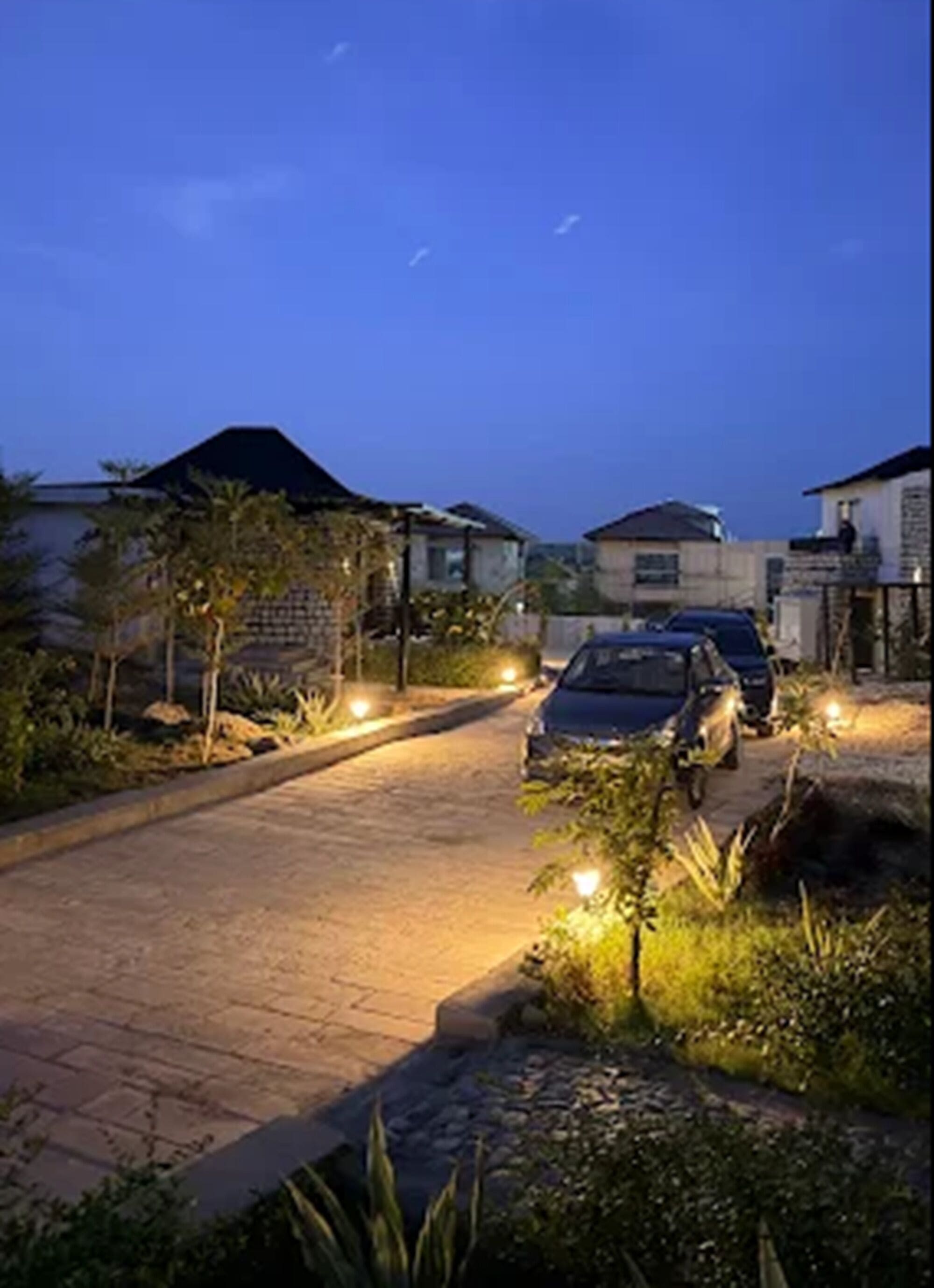 A residential street at dusk with houses on either side, a paved road, and a parked car.