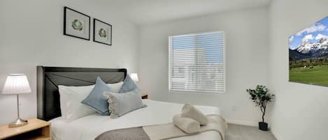 Ein Schlafzimmer mit einem Bett, Nachttischlampen, einem Fenster mit Jalousien, einem gerahmten Bild und einer Pflanze.