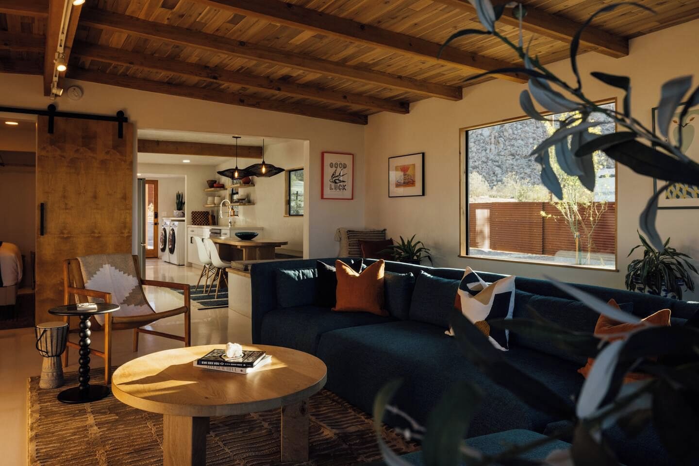A modern living room with a dark blue sofa, a round wooden coffee table, and a dining area with a kitchen in the background.