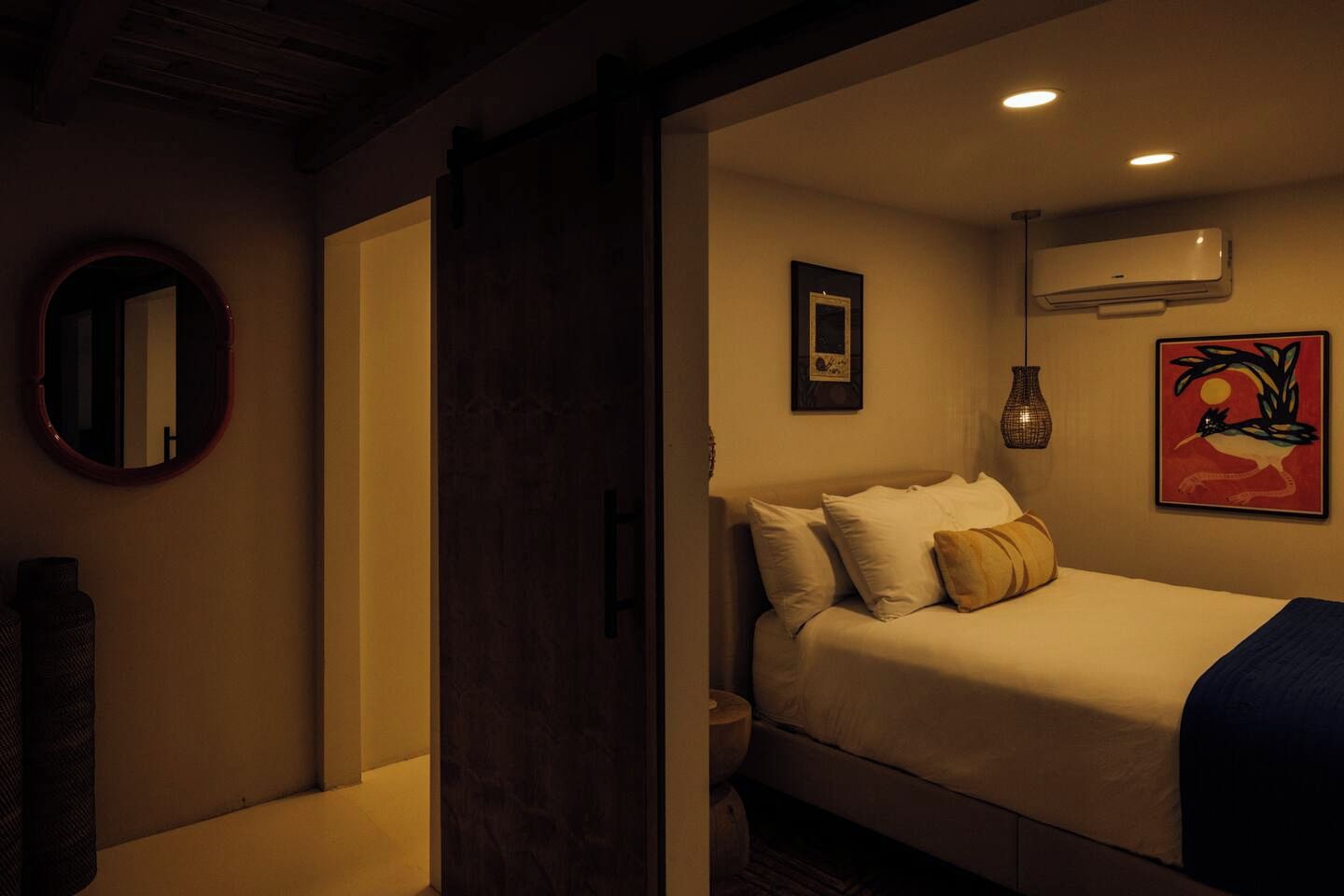 Un dormitorio con cama, almohadas, una mesita de noche, un espejo, un cuadro enmarcado, un equipo de aire acondicionado y un jarrón decorativo.