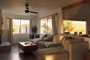 A living room with a sofa, a coffee table, and a ceiling fan. There are windows with curtains, a plant, and a view of the outdoors. - Black Rock Retreat - Desert Escape with Pool, Spa & Hammocks (Yucca Valley)
