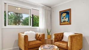 A room with two armchairs, a round coffee table, a painting, and a window with a view of trees. - Okere Bush Retreat - Sauna, Spa, Sleeps 8 (Okere Falls)