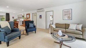 A living room with a sofa, armchair, and coffee table. There is a dining area with a table and chairs. The room has a white ceiling and walls, and a framed picture on the wall. - Okere Bush Retreat - Sauna, Spa, Sleeps 8 (Okere Falls)