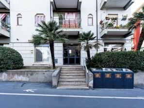 A multi-story residential building with a central entrance, a staircase leading to the entrance, and a row of potted plants on the balconies. - Nautilus by Interhome (Alassio)