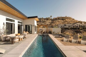 Un espace piscine extérieur moderne, doté de chaises longues et d’une pergola, entouré d’un paysage désertique et de collines.