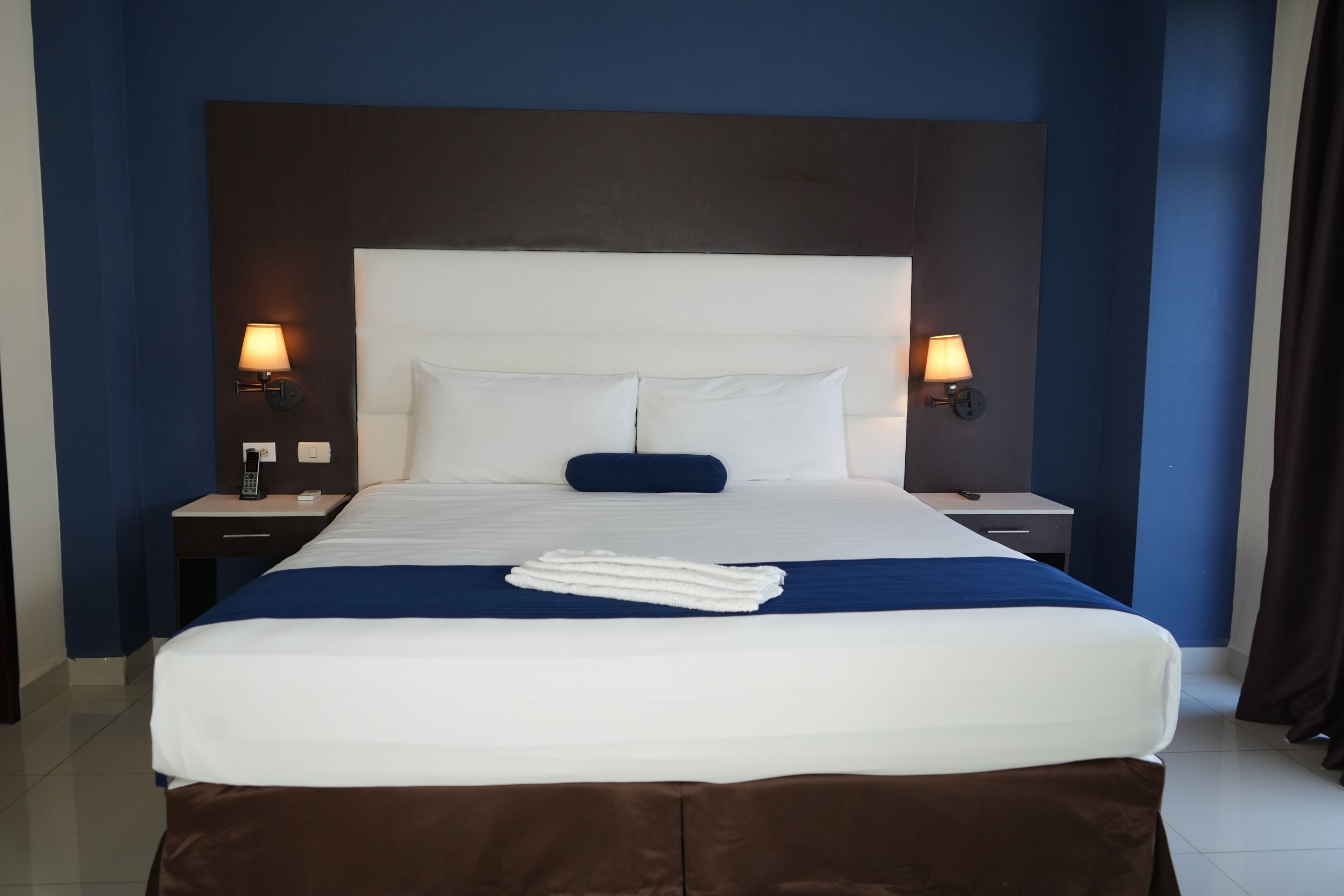 A hotel room with a large bed, two bedside tables, a headboard, and two wall-mounted lamps.
