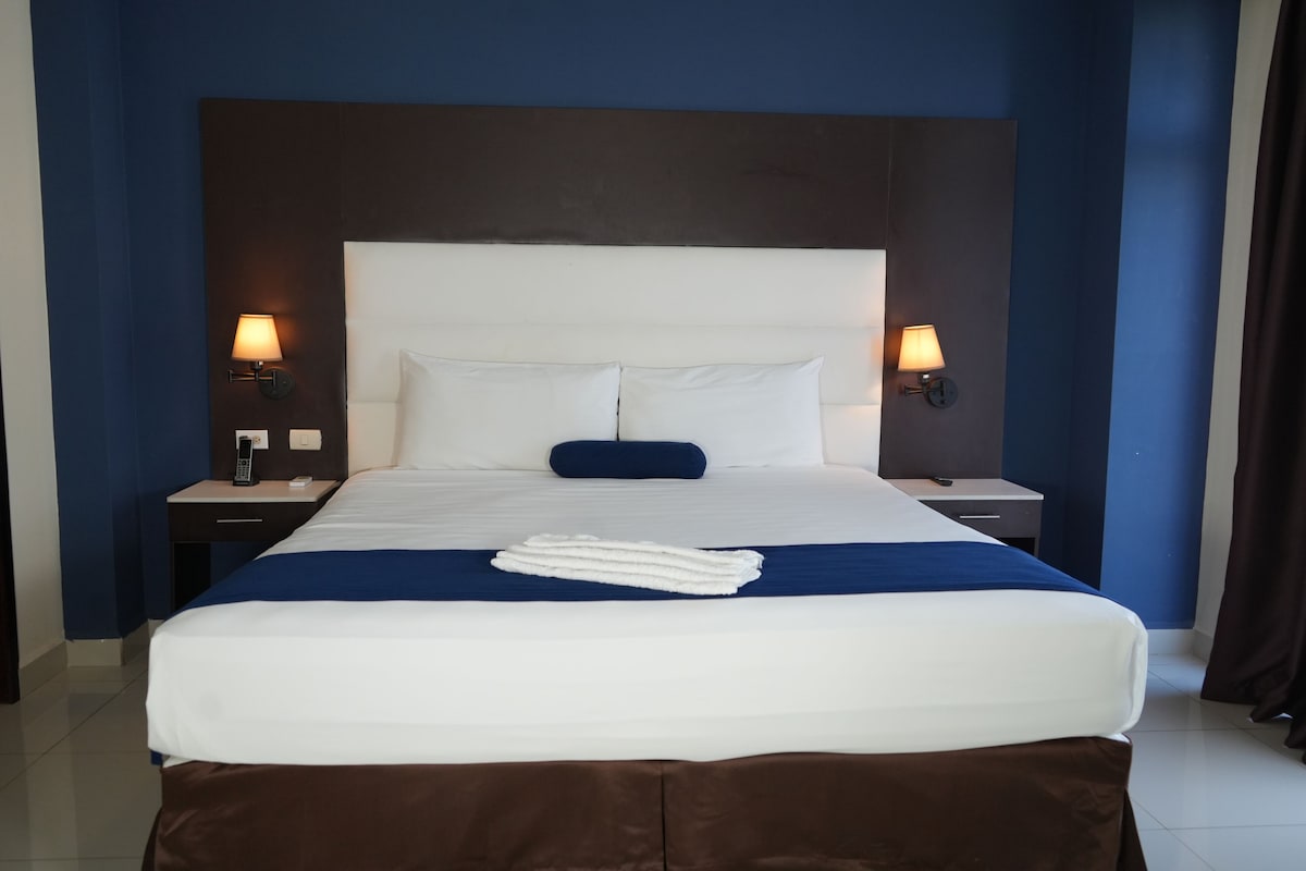 A hotel room with a large bed, two bedside tables, a headboard, and two wall-mounted lamps.