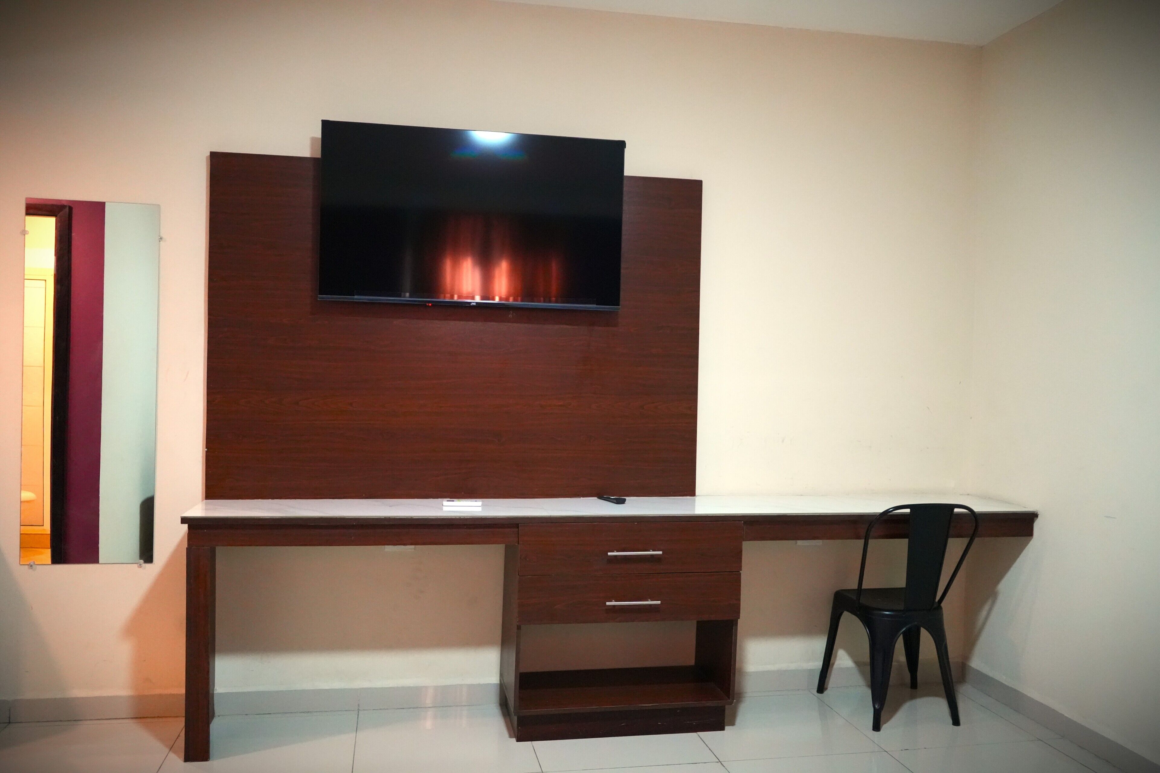 A room with a wooden desk, a television mounted on the wall, a chair, and a door with a mirror.