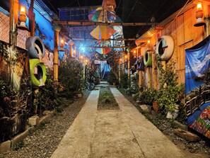 A narrow pathway flanked by walls adorned with tires and hanging lanterns, leading to a covered area with a view of a body of water.