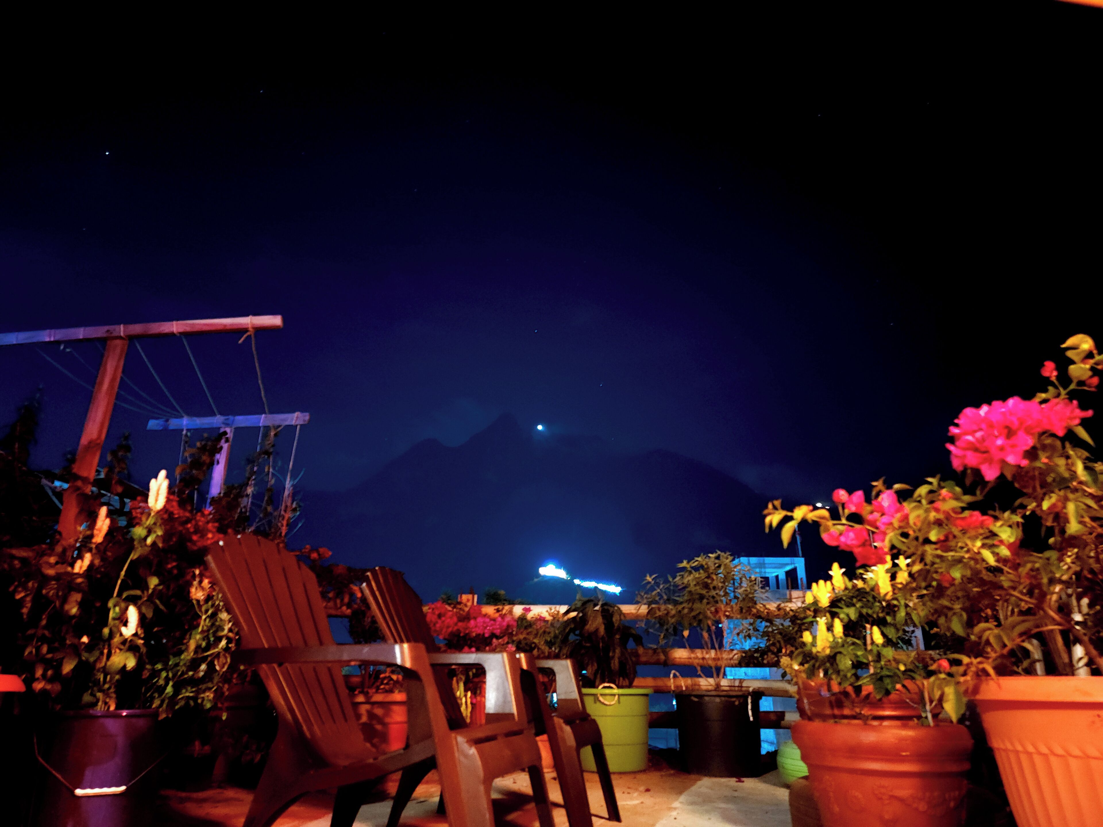 A patio with chairs and potted plants, a mountain in the background, and a building with lights.