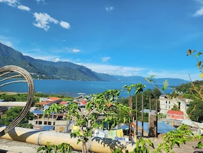 A coastal town with a view of the sea and mountains, featuring buildings with red roofs and a large metal sculpture.