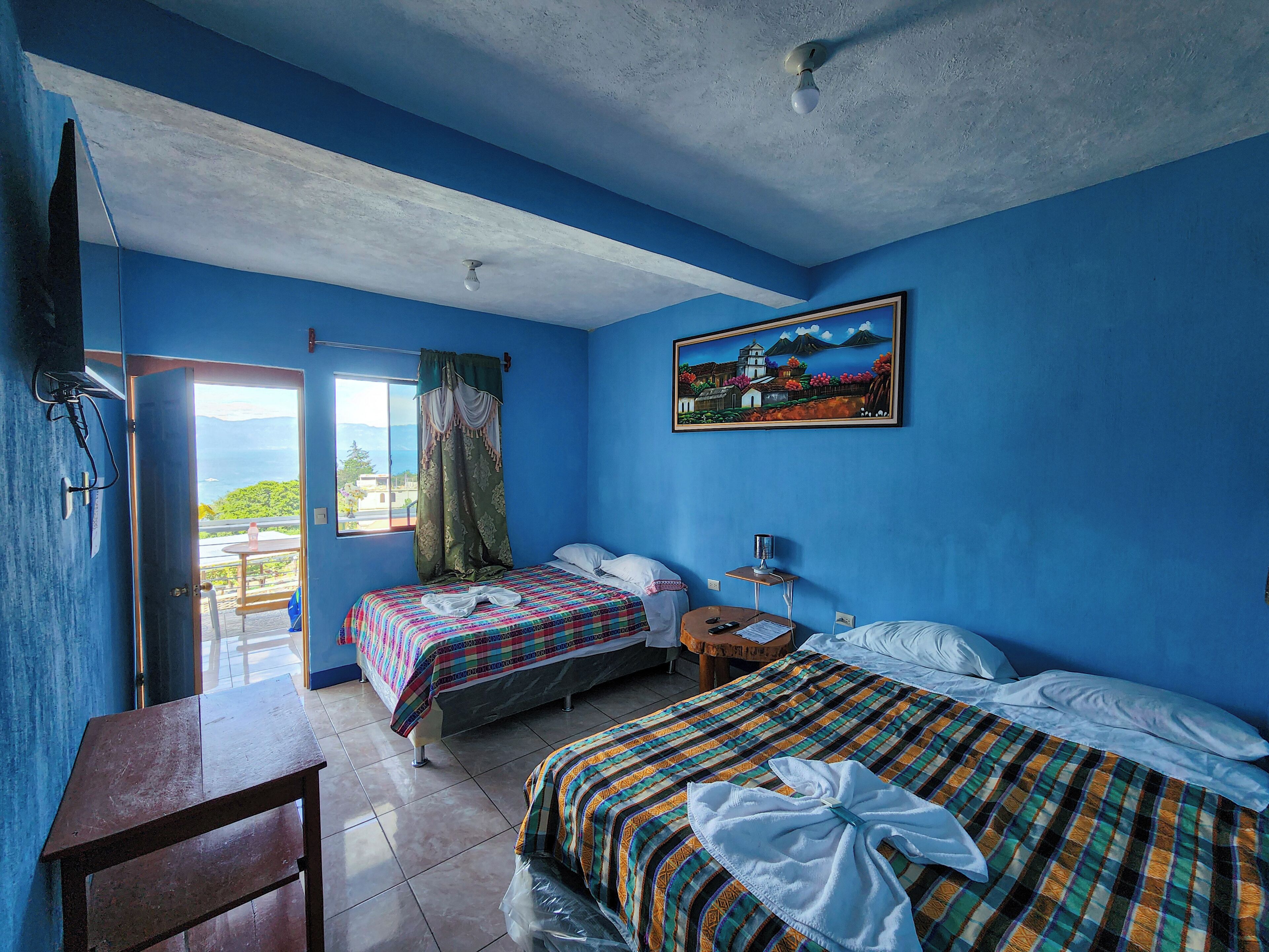 Habitación con dos camas, un escritorio, una silla, un ventanal con cortinas, un cuadro en la pared y un ventilador de techo.