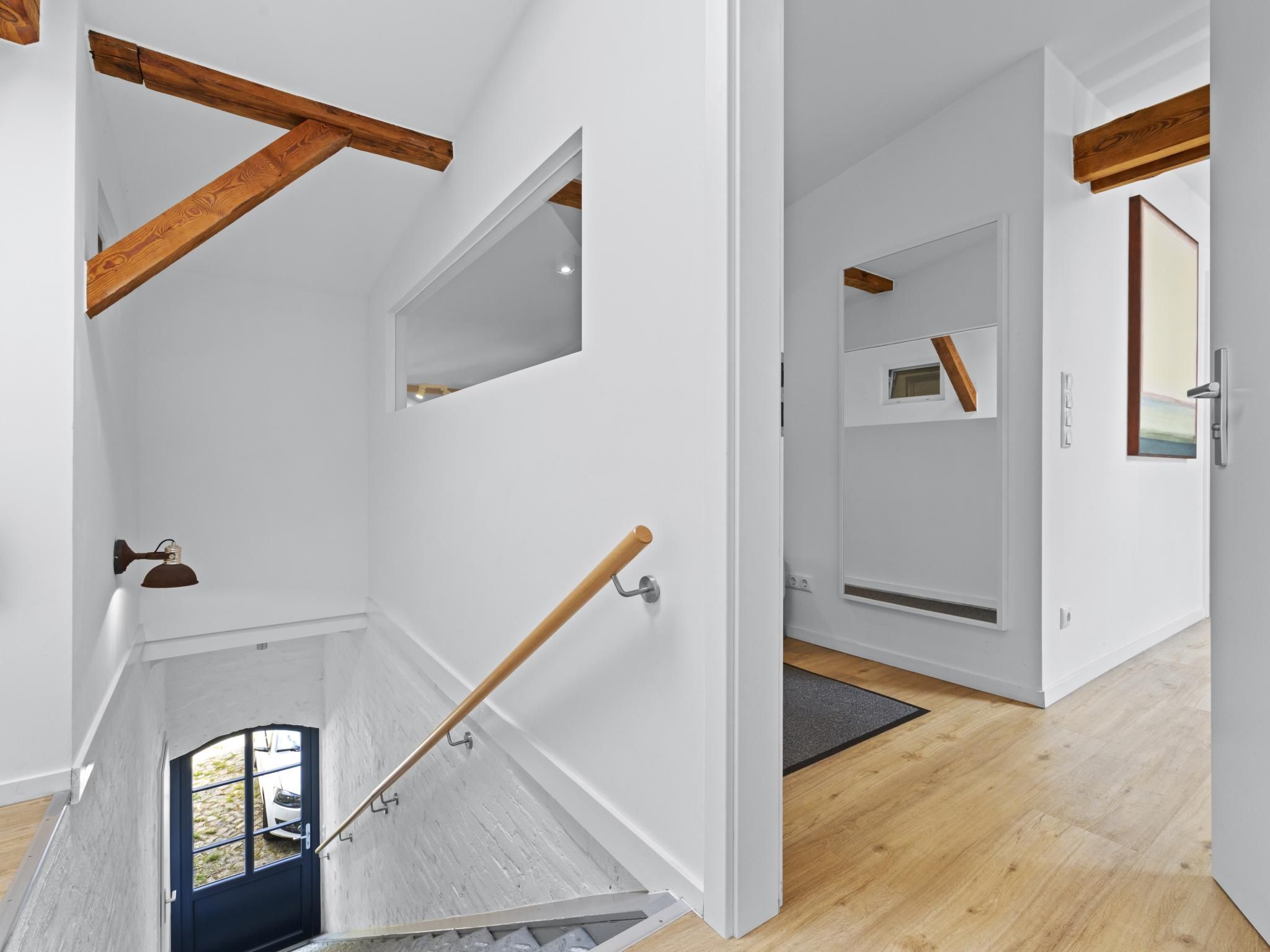 Un interior moderno con una escalera de madera, paredes blancas y vistas a través de una puerta.