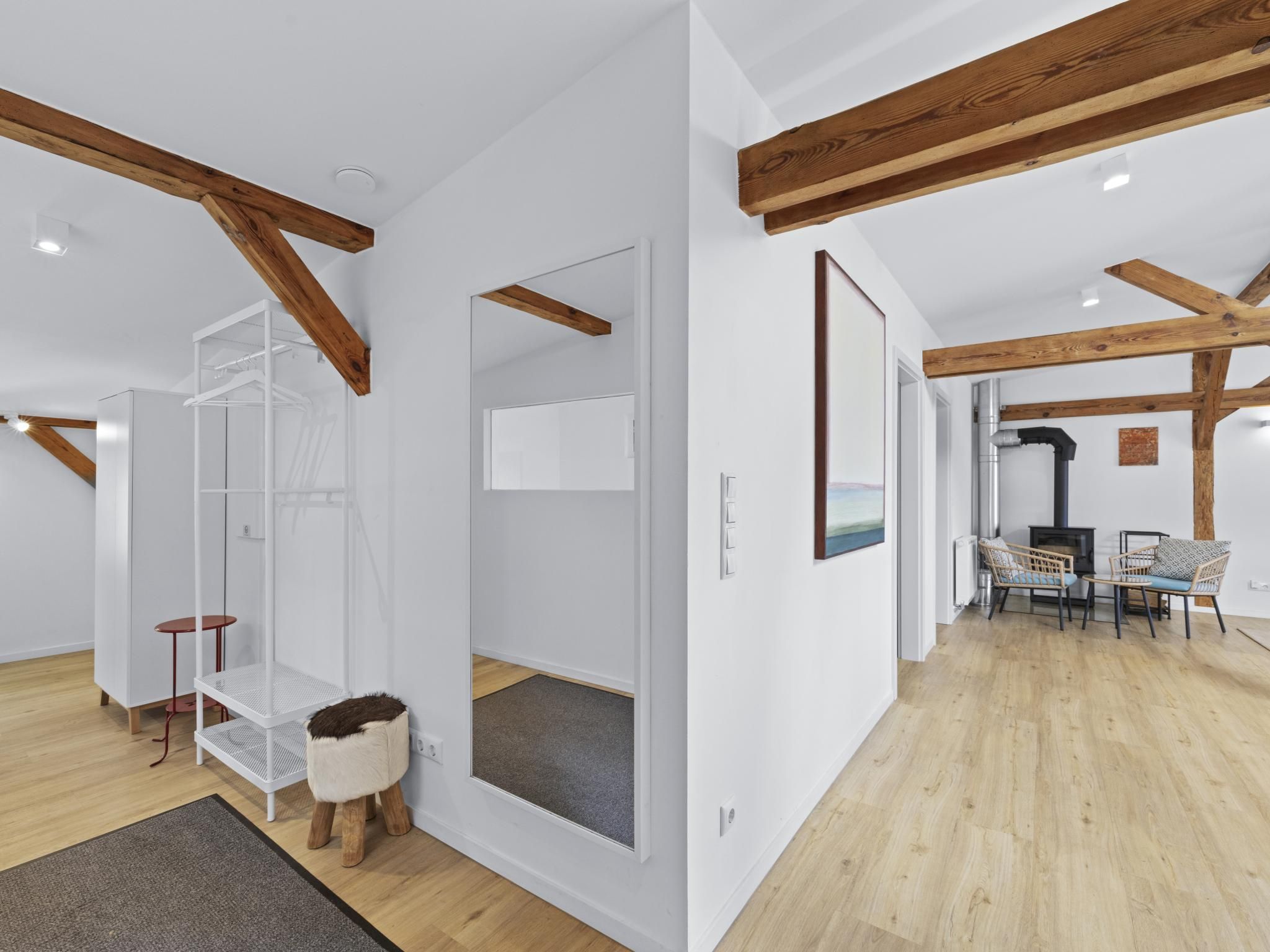 Ein modernes Interieur mit freiliegenden Holzbalken, einem weißen Kleiderschrank, einem kleinen Hocker und einem Essbereich mit Korbstühlen.