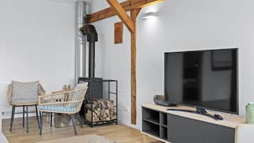 Ett modernt vardagsrum med TV, synliga träbjälkar, en öppen spis och en matplats med stolar i rotting.