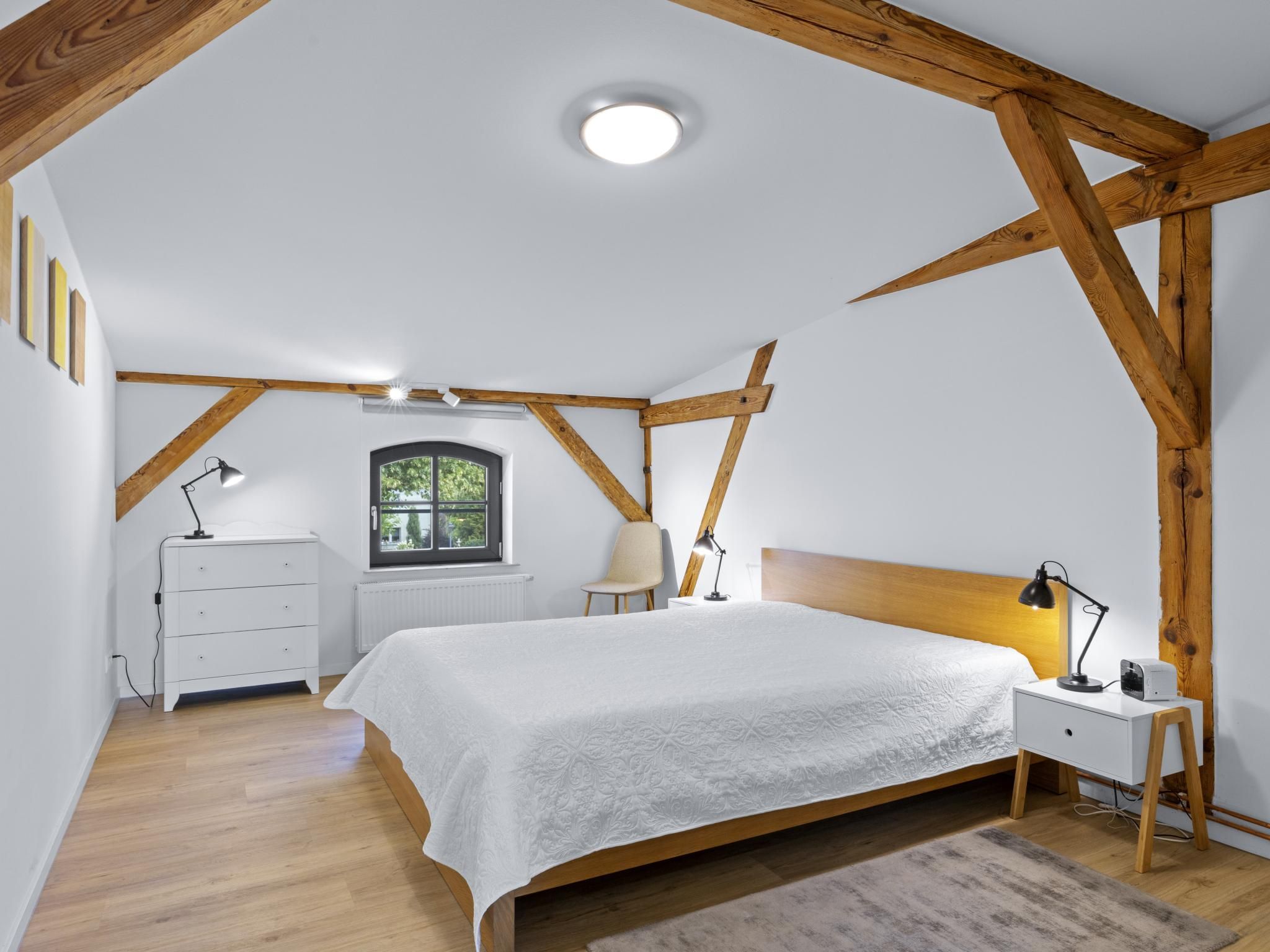 Ein Schlafzimmer mit geneigter Decke, Holzbalken, einem Bett mit weißer Bettdecke, Nachttischlampen, einem Nachttisch, einer Kommode und einem Fenster mit Blick auf Grünflächen.