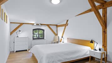Ein Schlafzimmer mit geneigter Decke, Holzbalken, einem Bett mit weißer Bettdecke, Nachttischlampen, einem Nachttisch, einer Kommode und einem Fenster mit Blick auf Grünflächen.