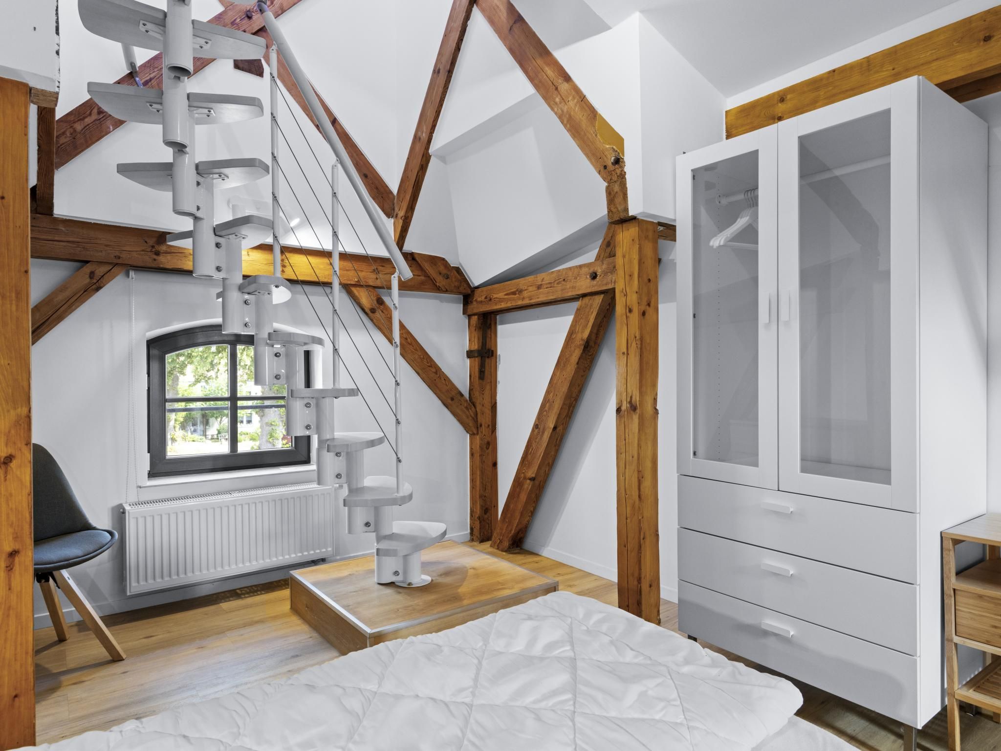 Een kamer met een wit kastje, een raam met uitzicht, een wit bed en een houten plafondconstructie.