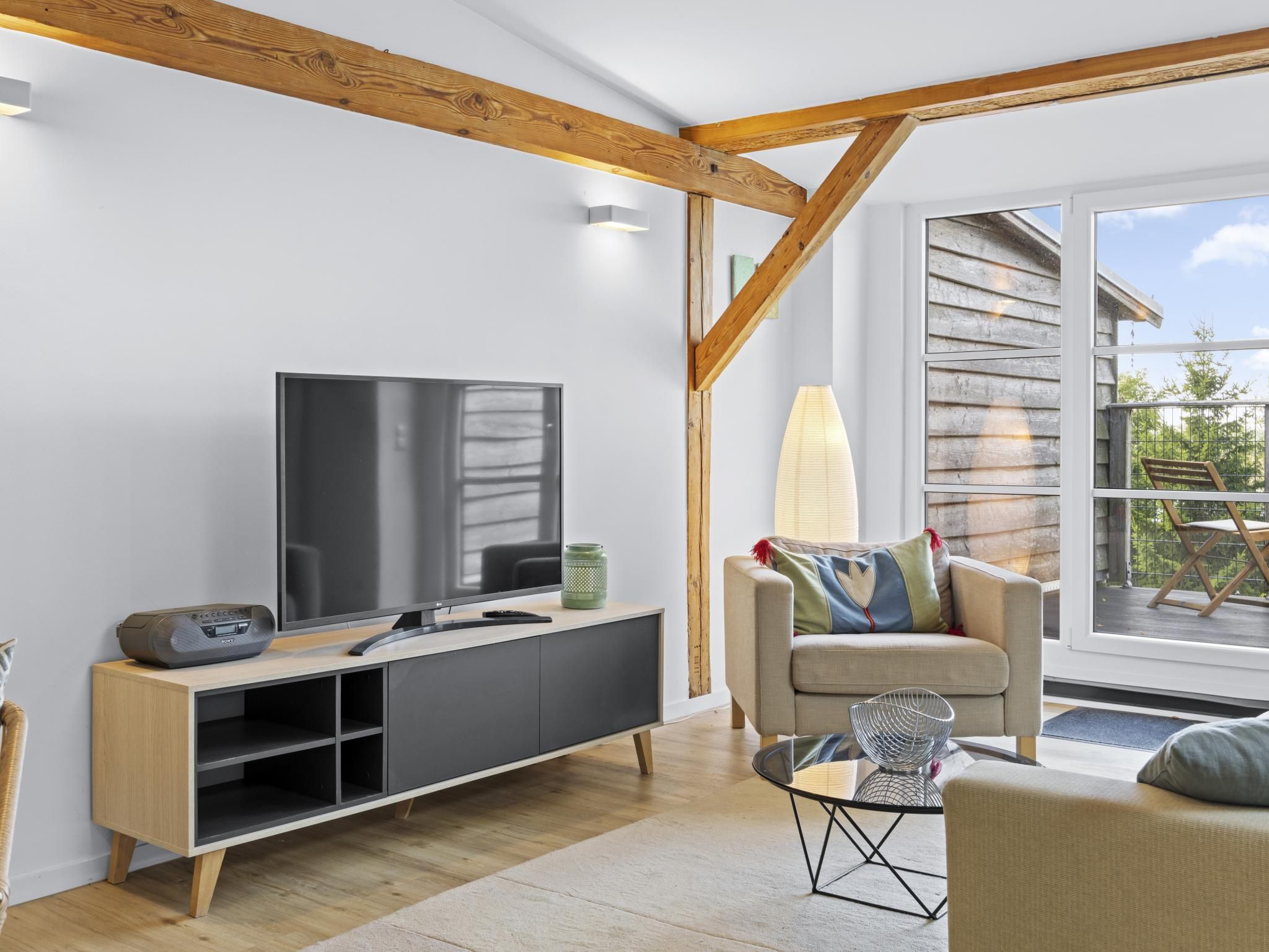 Ein modernes Wohnzimmer mit Flachbildfernseher, Holzbalken, einer Couch und Blick ins Freie.