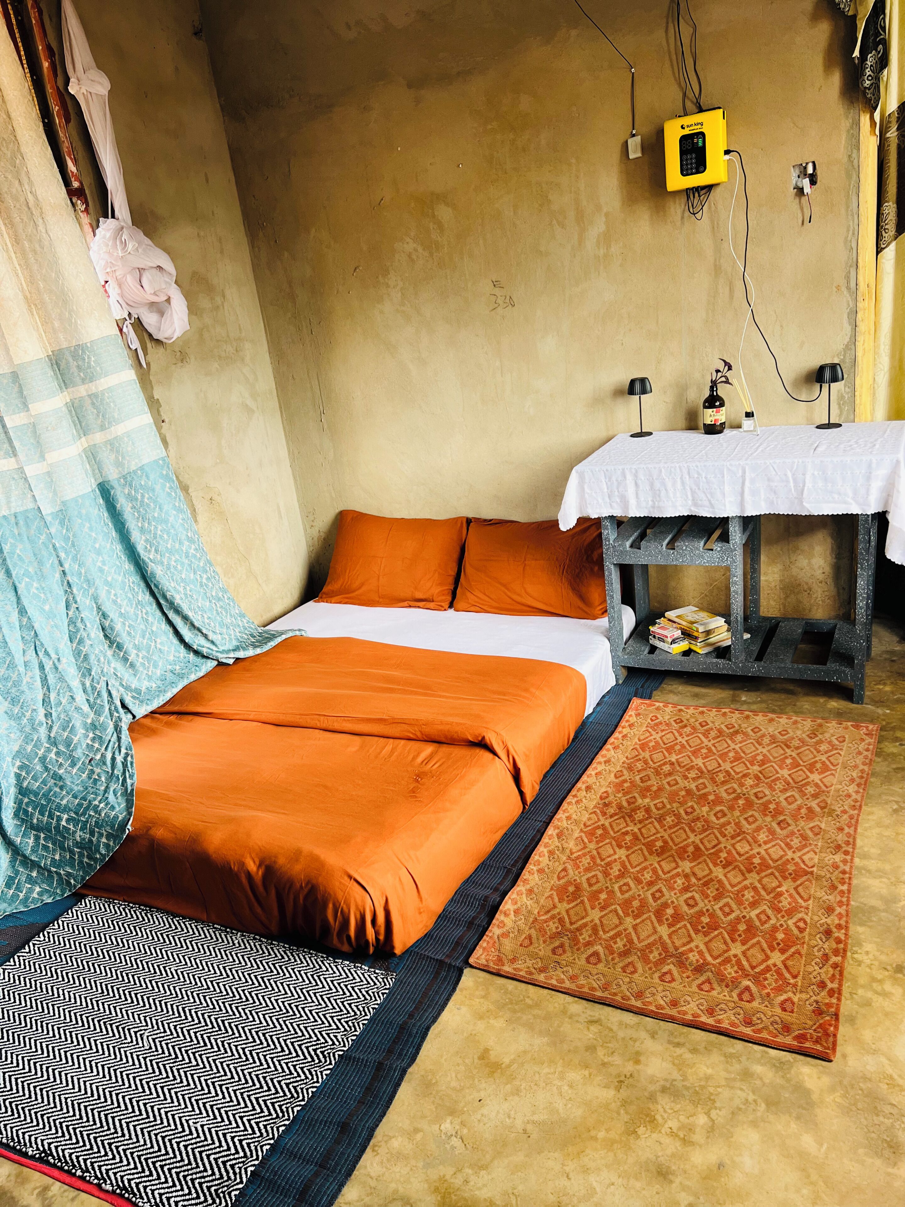 A room with a bed, a small table with a white tablecloth, a patterned rug, and a wall-mounted device.