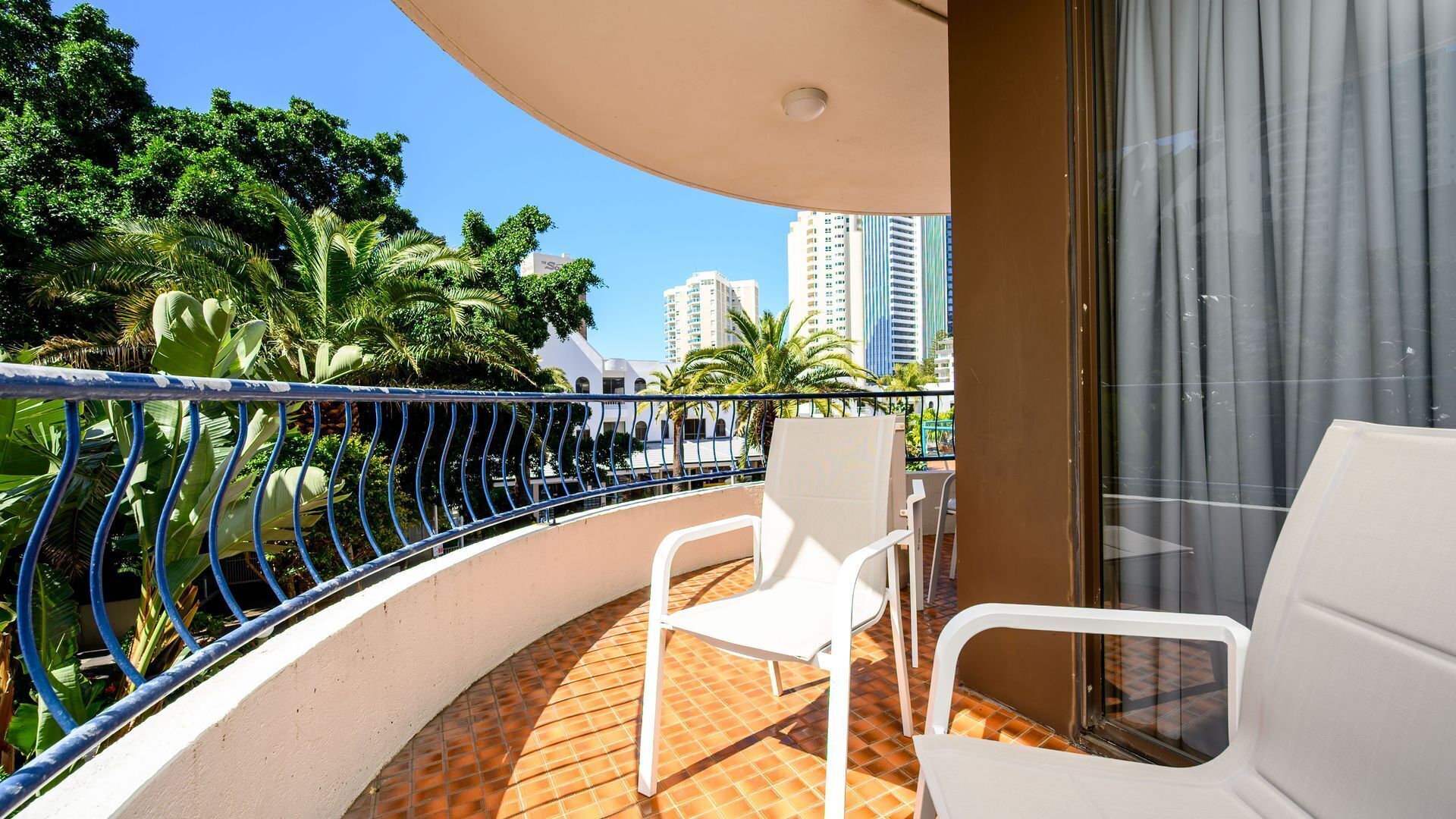 A balcony with a white chair, a glass door, and a view of tall buildings and greenery.