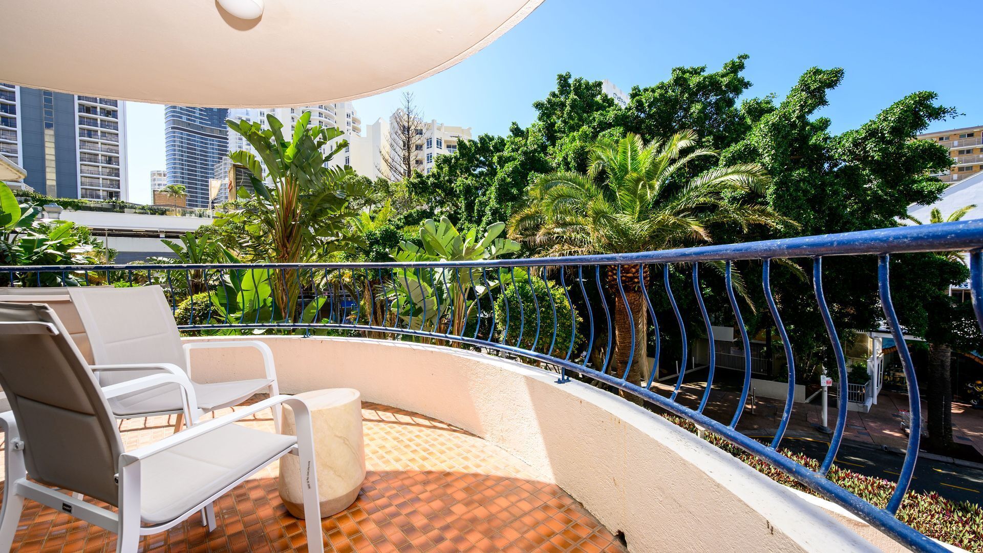 A balcony with white chairs and a small table, surrounded by a blue railing, overlooking a cityscape with buildings and lush greenery.