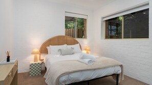 Een slaapkamer met een bed, twee bedlampen, een nachtkastje met ruitdessin en een raam met uitzicht op groen.