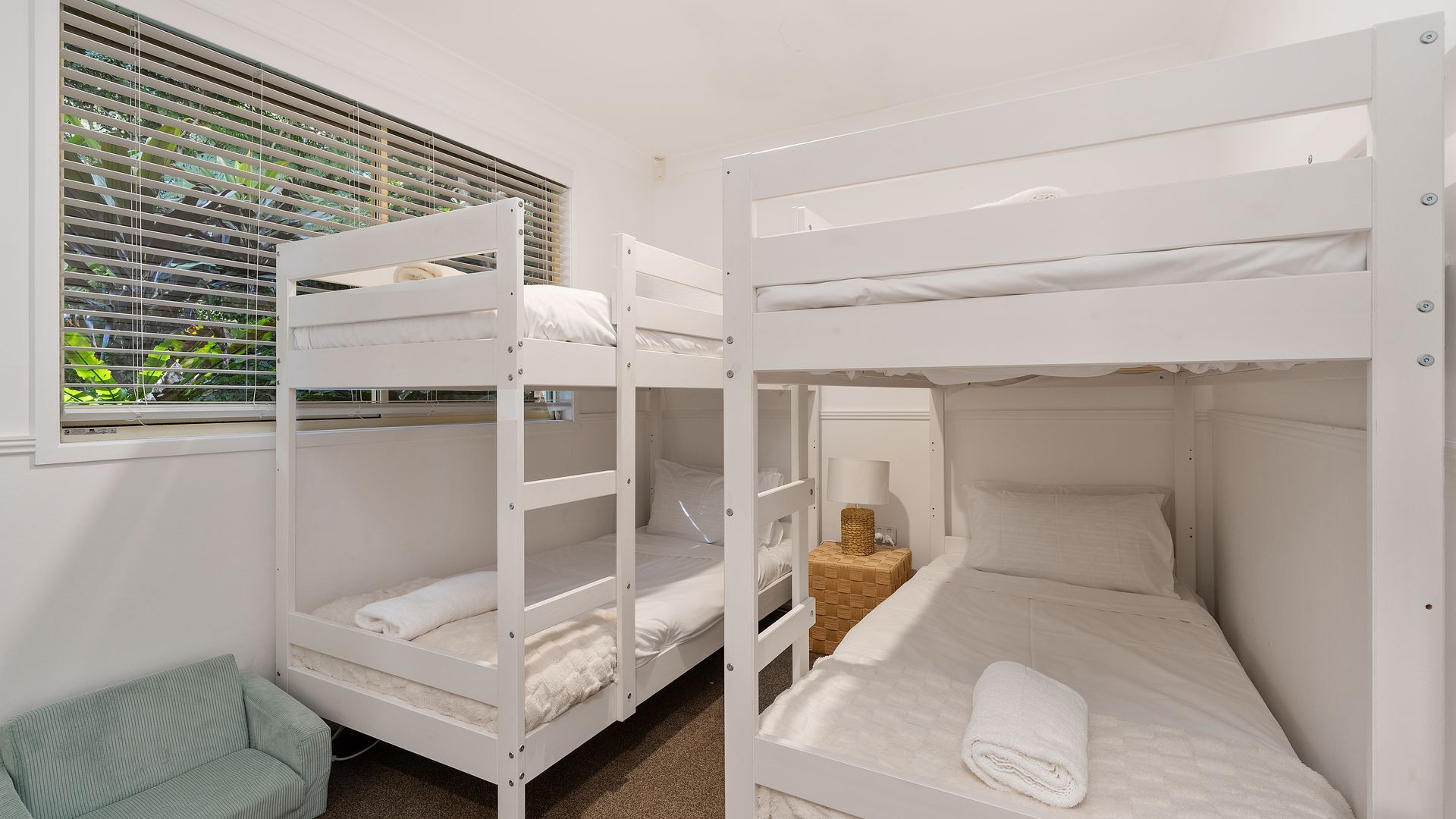 A room with bunk beds, a window with blinds, a bedside table with a lamp, and a small chair.
