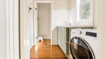 A laundry room with a washing machine, a small stool, and a window with a view of trees.