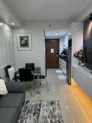A modern living room with a sofa, a dining table with chairs, a kitchen area with a refrigerator, and a TV mounted on the wall. - Cozy Scandinavian-inspired Condo With Balcony Facing SM Mall of Asia (Pasay)